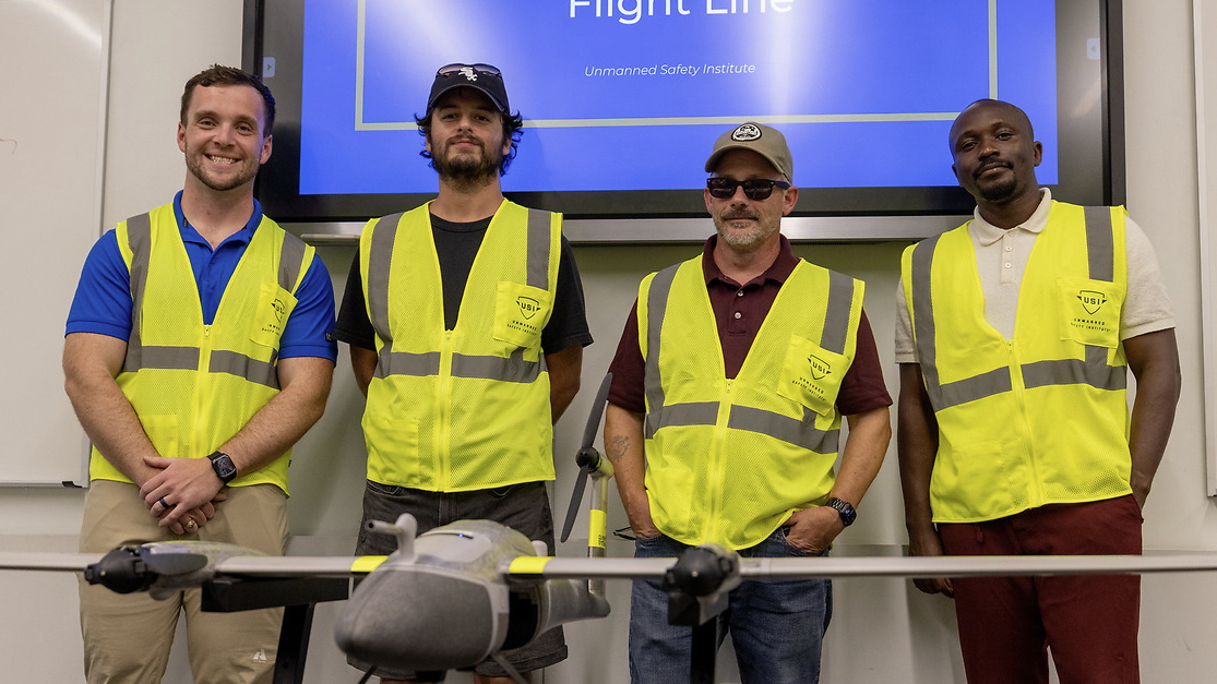 Four people wearing high-visibility safety vests stand behind a fixed-wing drone displayed on a table in a classroom or training room. A presentation screen is visible in the background.
