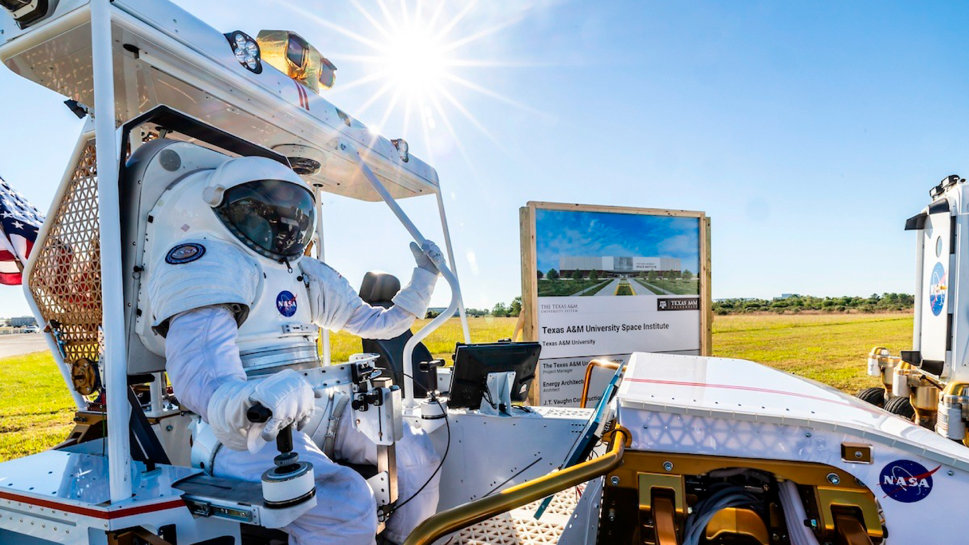 An astronaut in a NASA spacesuit sits in a lunar rover prototype during a groundbreaking event for the Texas A&M University Space Institute. A rendering of the future facility is visible in the background.