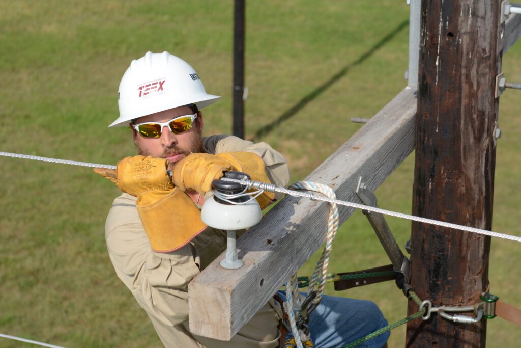 A utility worker wearing a hard hat and gloves is working on a wooden utility pole with wires and equipment.
