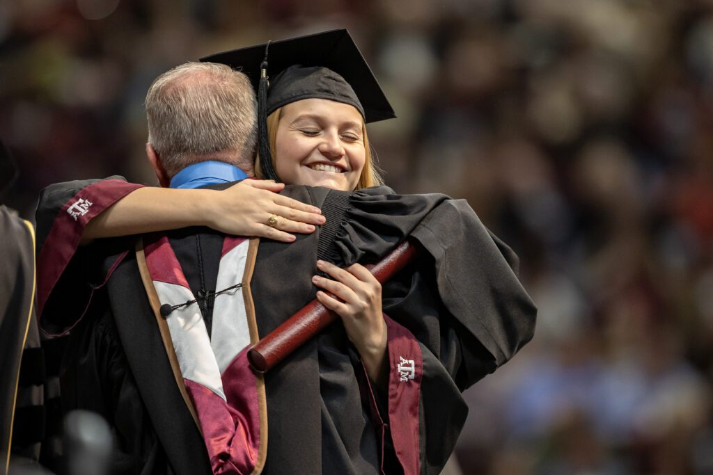 A graduate in a cap and gown embraces a person in academic regalia, smiling joyfully while holding a diploma.