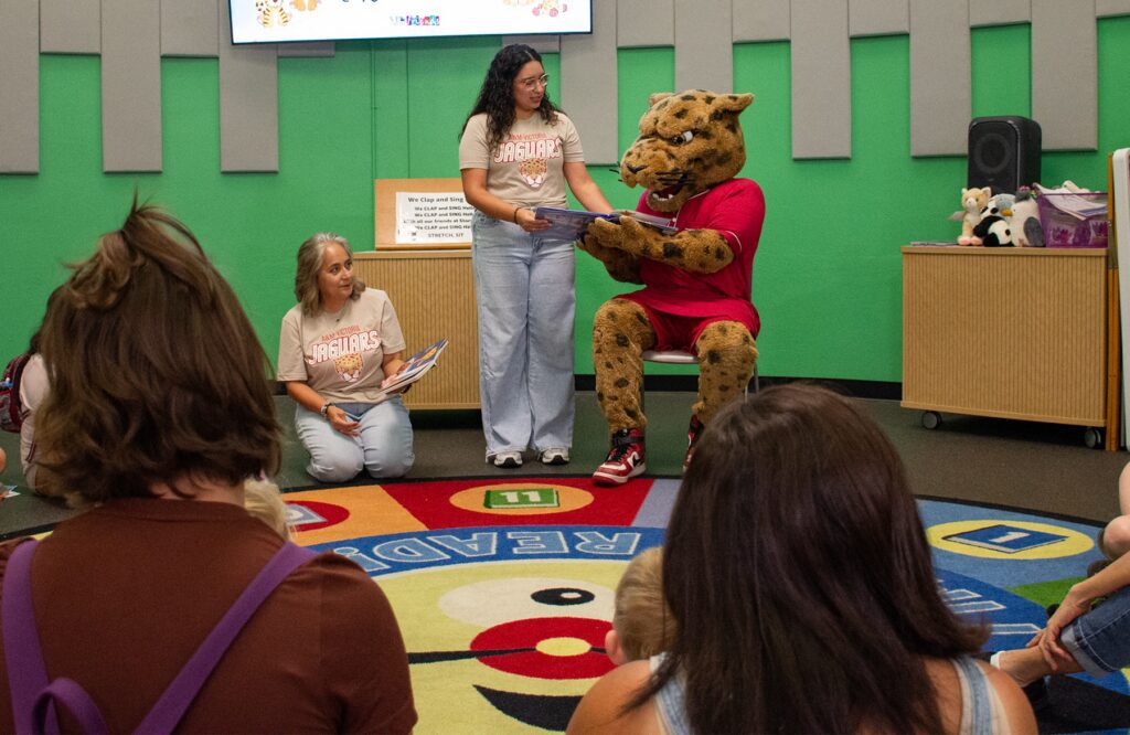 School mascot reads a book to children during a storytime event as two adults assist.